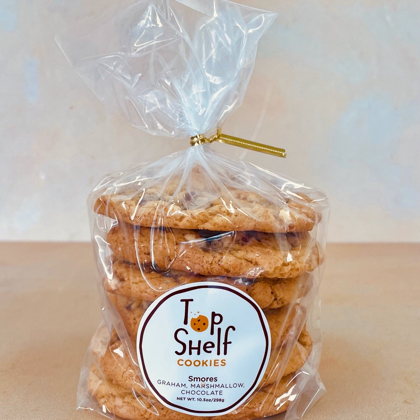A pack of S'Mores cookies wrapped in a transparent plastic bag with a gold-colored twist tie on top. The packaging has a brown label with the brand 'Top Shelf Cookies' and the product name 'S'Mores'.