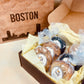 A box of assorted cookies from Top Shelf Cookies, packaged with individual clear wrapping and a label indicating the flavor, placed inside a cardboard box with a 'BOSTON' printed on it.