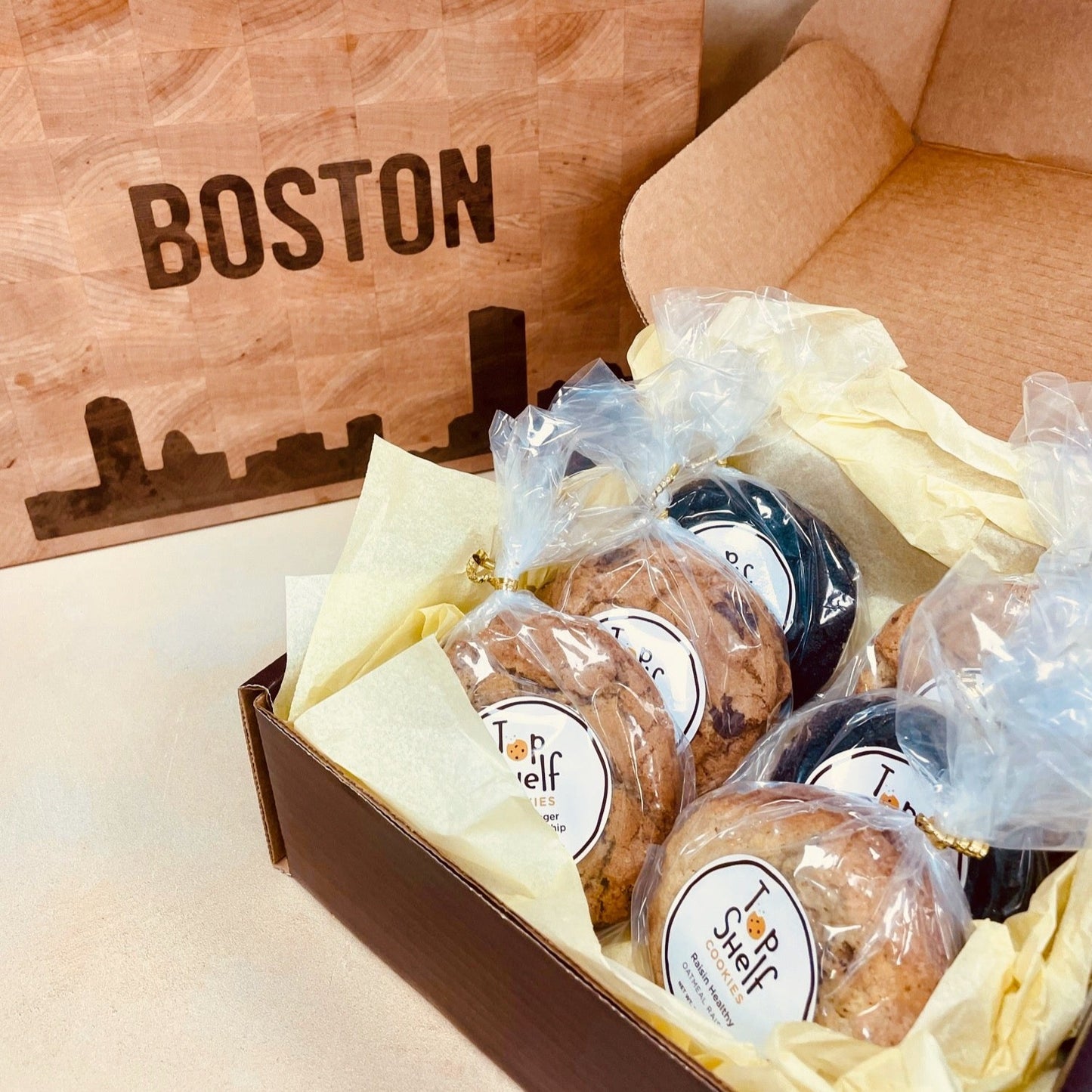 A box of assorted cookies from Top Shelf Cookies, packaged with individual clear wrapping and a label indicating the flavor, placed inside a cardboard box with a 'BOSTON' printed on it.